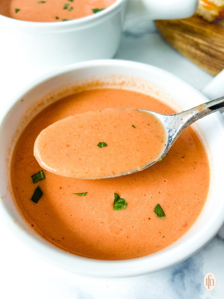 Spoon lifting tomato soup out of a bowl with pieces of basil on top.