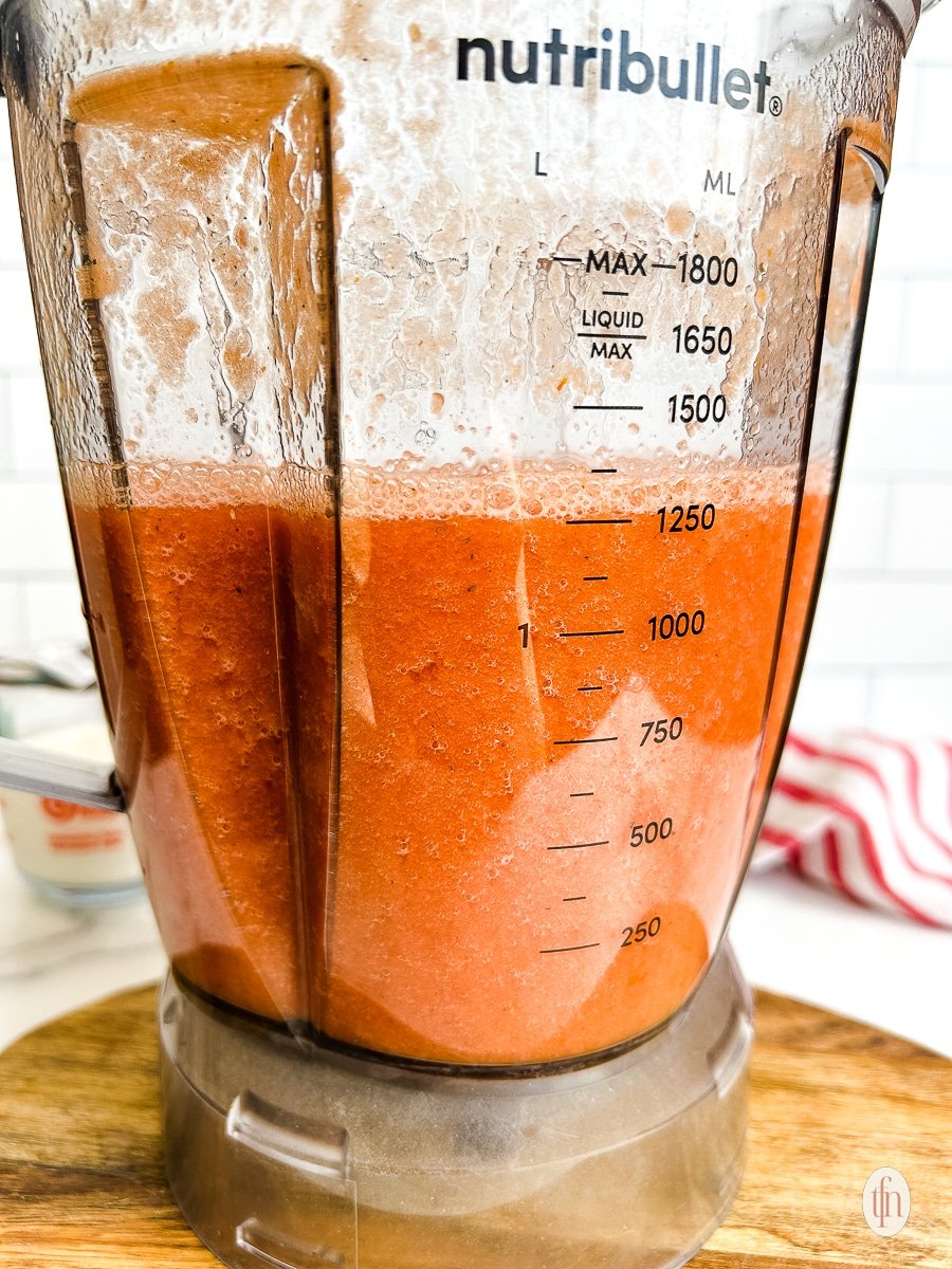 Blender with tomato soup on a cutting board.