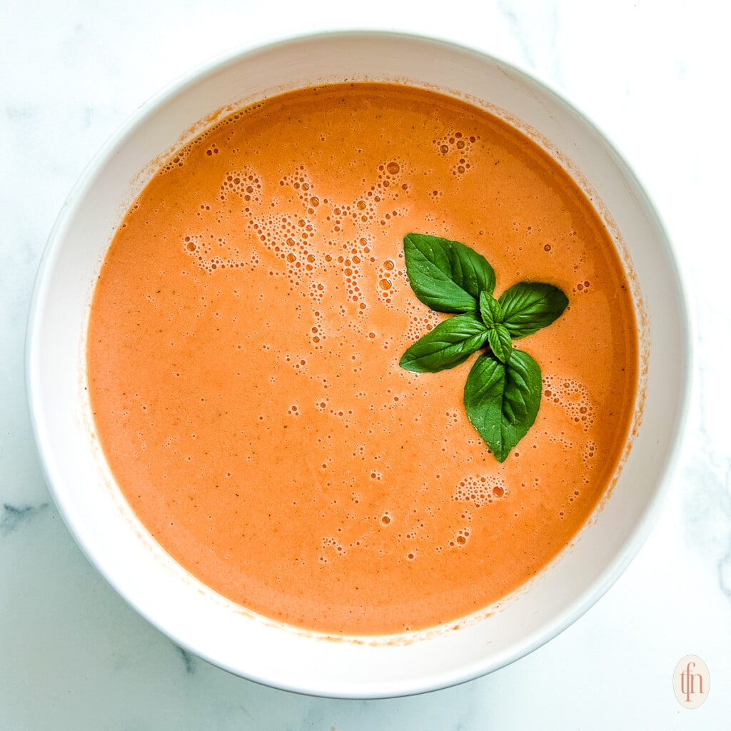 A white bowl filled with tomato soup and a sprig of basil on top.