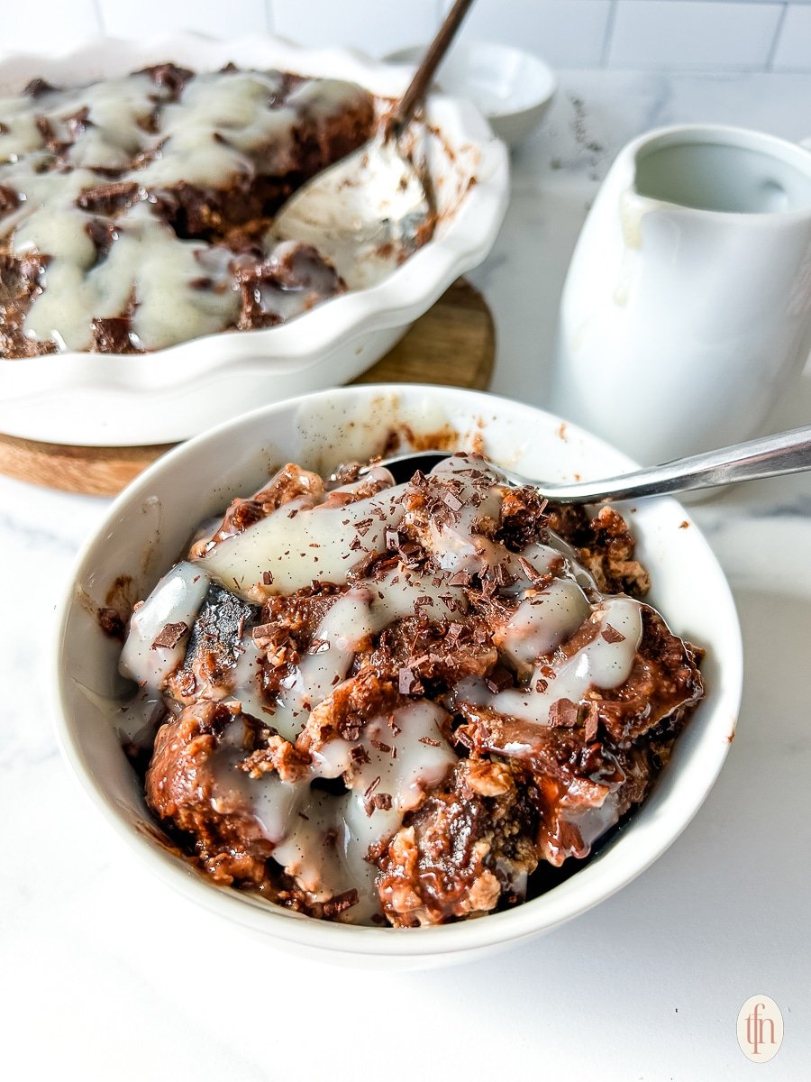 Chocolate bread pudding in a white bowl with vanilla cream sauce drizzled over the top.