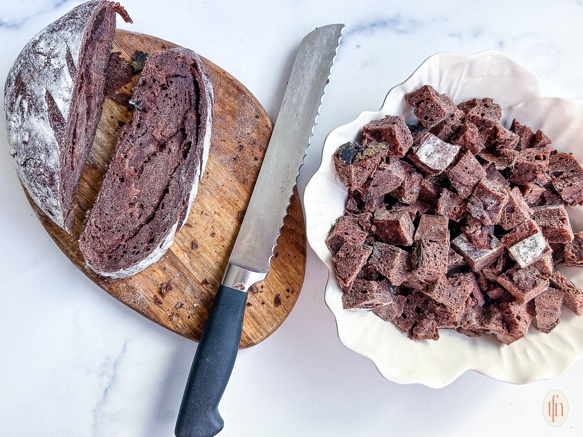 Slicing chocolate sourdough bread into cubes with a long serrated knife on a cutting board.