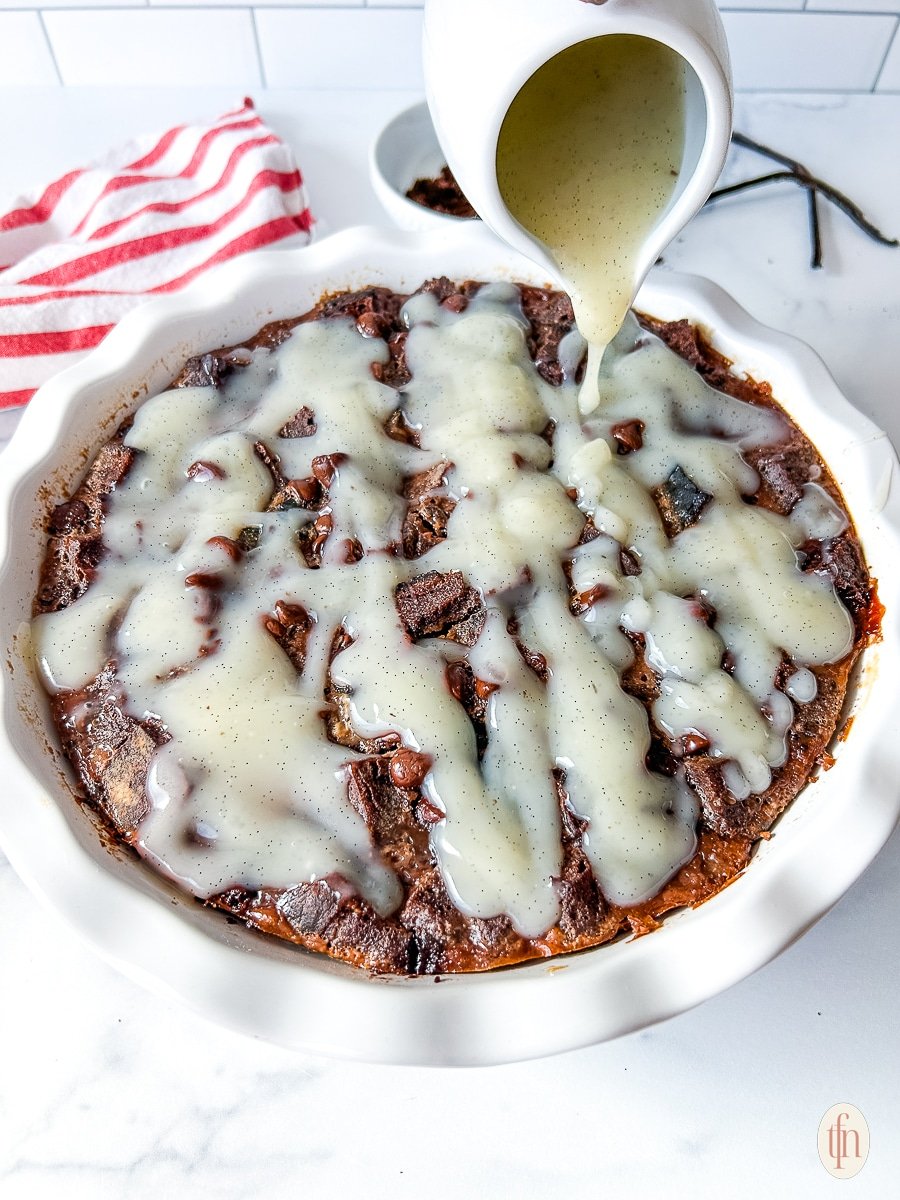 Drizzling vanilla cream sauce over bread pudding in a white pie dish.