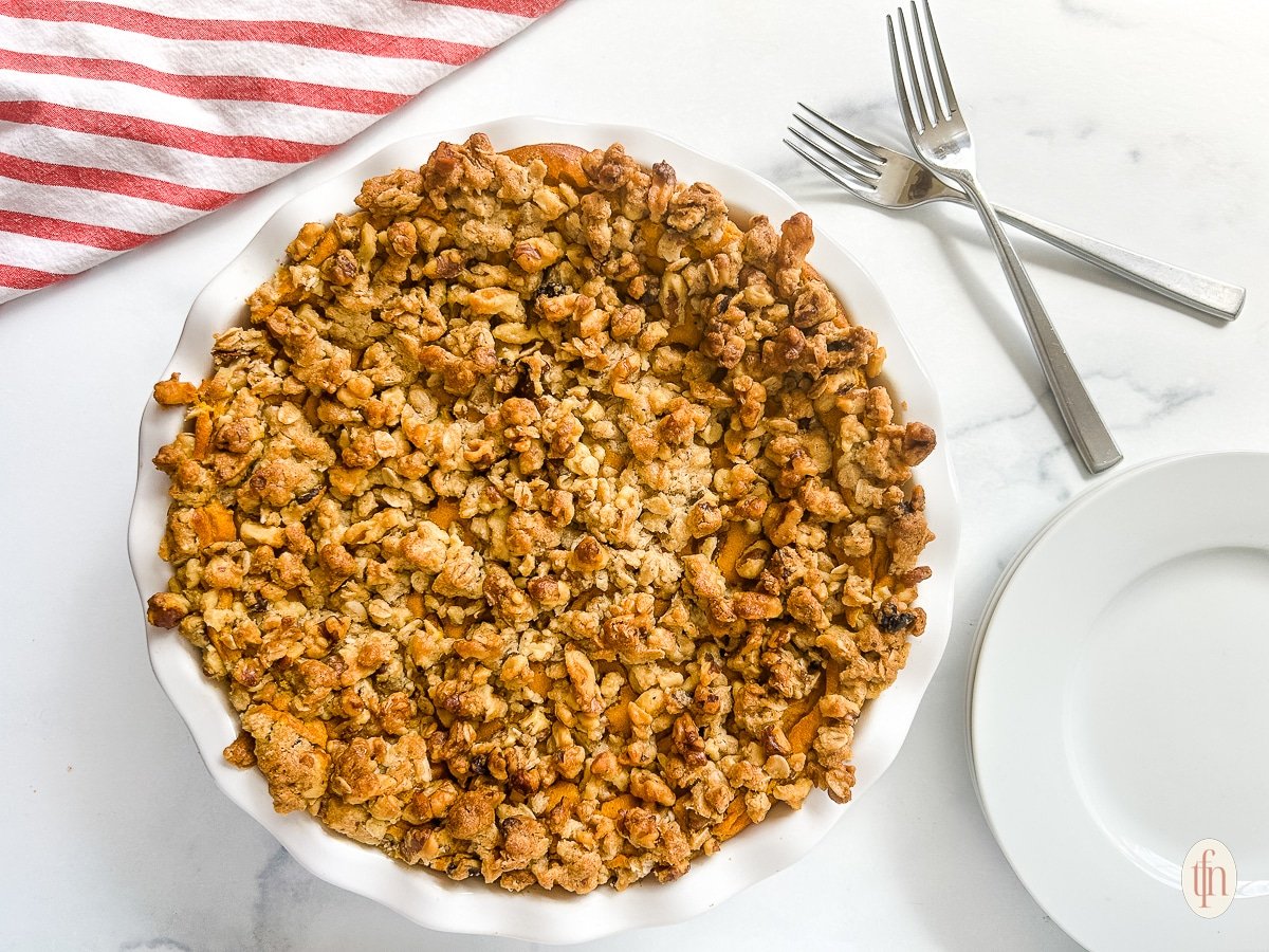 A whole pie with crumble on top next to an empty plate and two crossed forks on a white background.