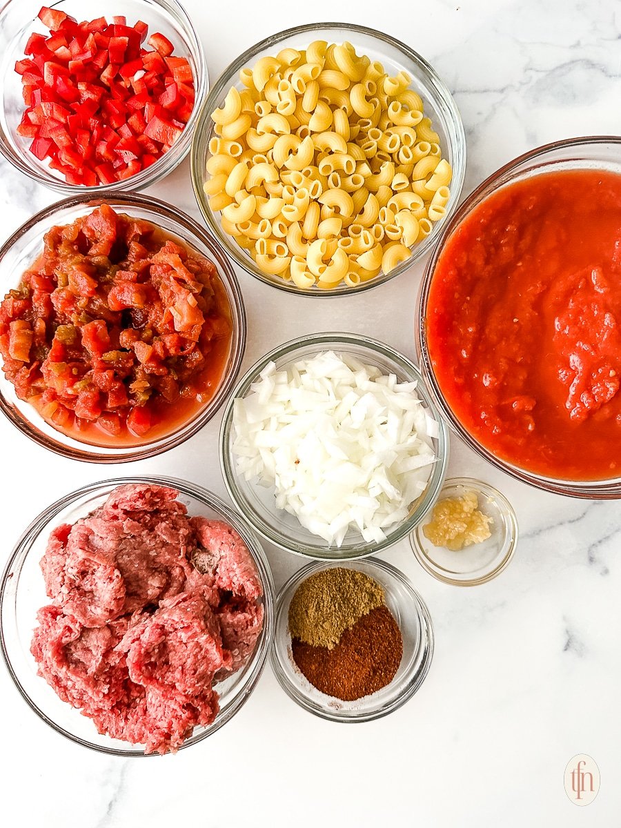 Small bowls with uncooked macaroni, ground beef, tomato puree, and other ingredients for Instant Pot goulash recipe.
