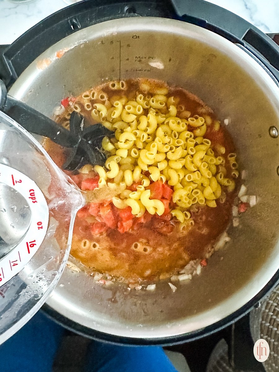 Adding hot water to a large pot of elbow macaroni in cooking liquid.