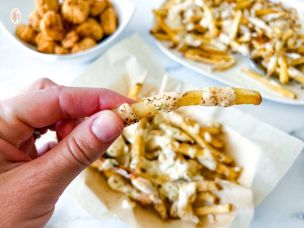 A hand holding up a French fry drizzled with white sauce over a plate of other fries.