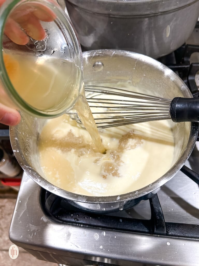 A cup of chicken broth being poured into bechamel sauce.