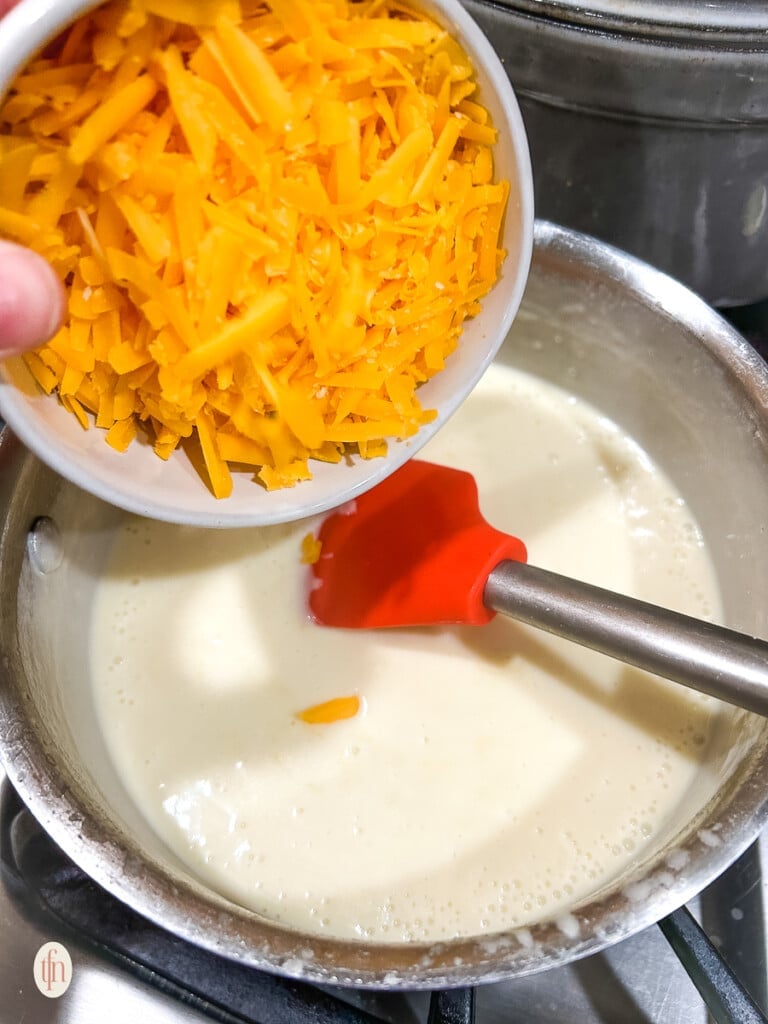 Hand adding a bowl of shredded cheese to a bechamel sauce.