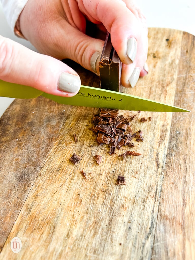Shaving chocolate with a knife on a cutting board.