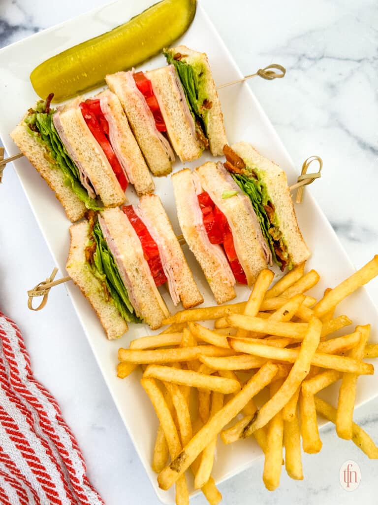 Four slices of a chicken club sandwich with sticks and fries on a white plate.