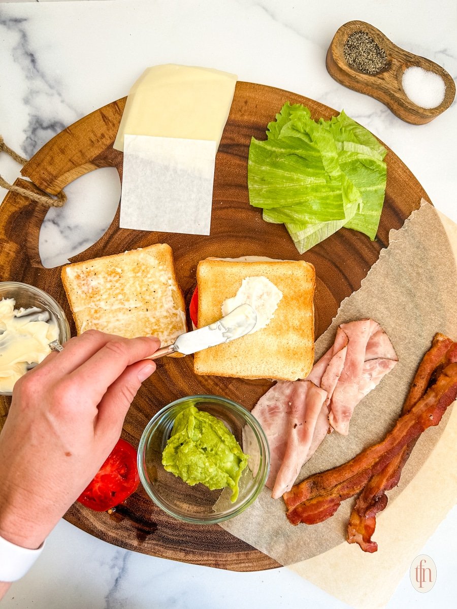 Spreading sauce on bread with prepared ingredients for chicken club sandwich on a round wood board.