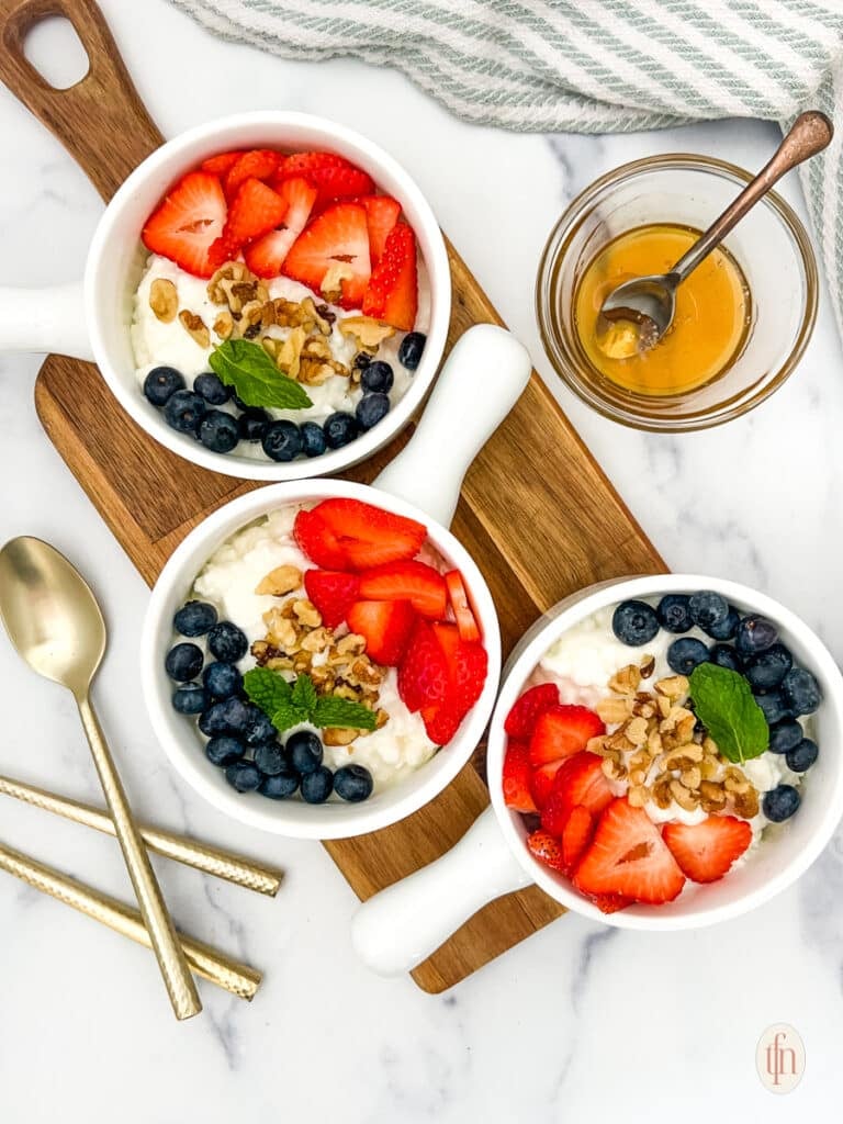 Three bowls of cottage cheese and fruit on a wooden board with spoon and a glass of honey.