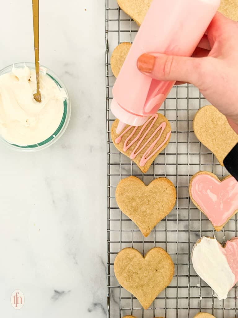Heart cookies being decorated on a cooling rack with a bowl of frosting.