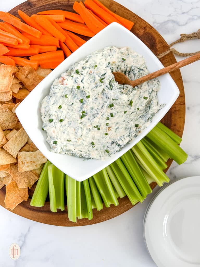 Knorr spinach dip on a serving rectangular bowl with a wooden spoon. Served on top of a round wooden board with crackers and vegetables.