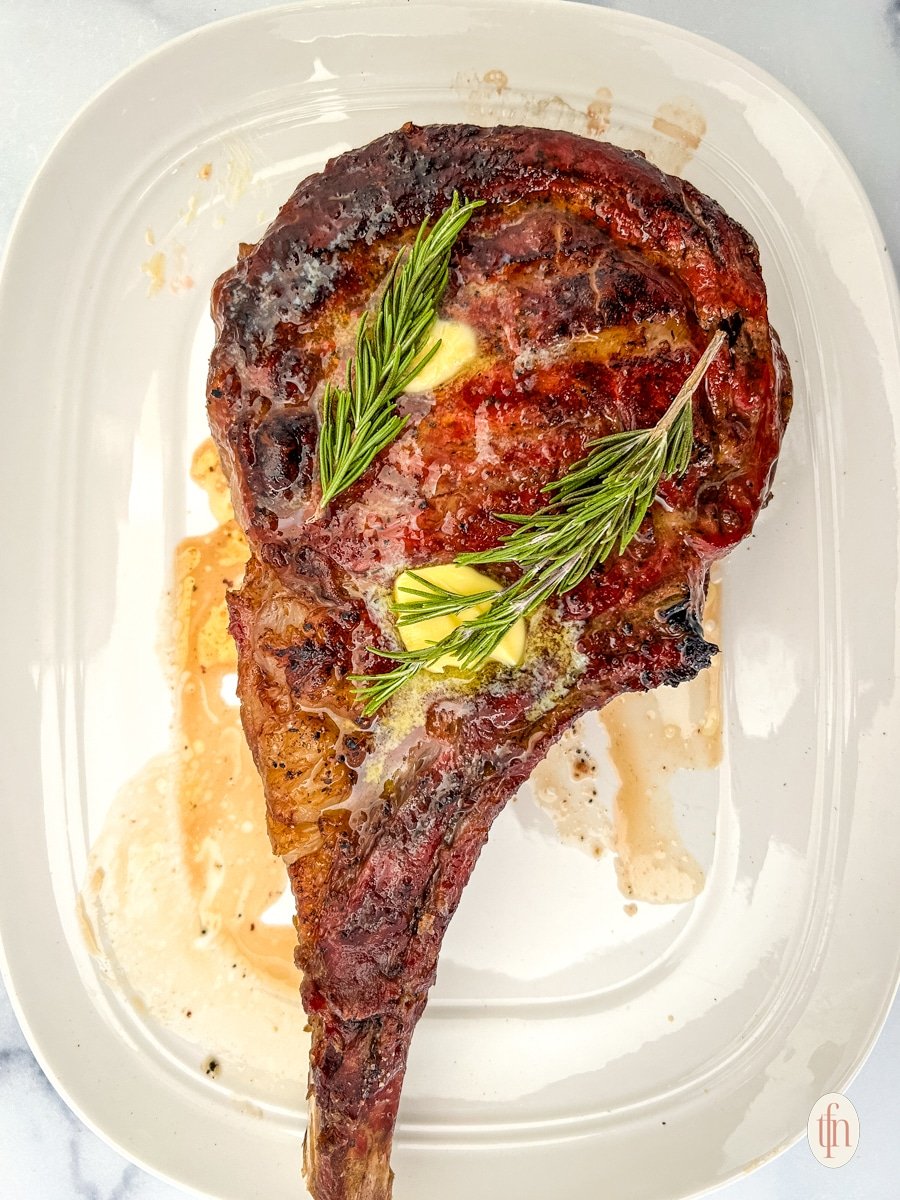 Perfectly cooked cowboy ribeye on a white plate with rosemary and butter on top.