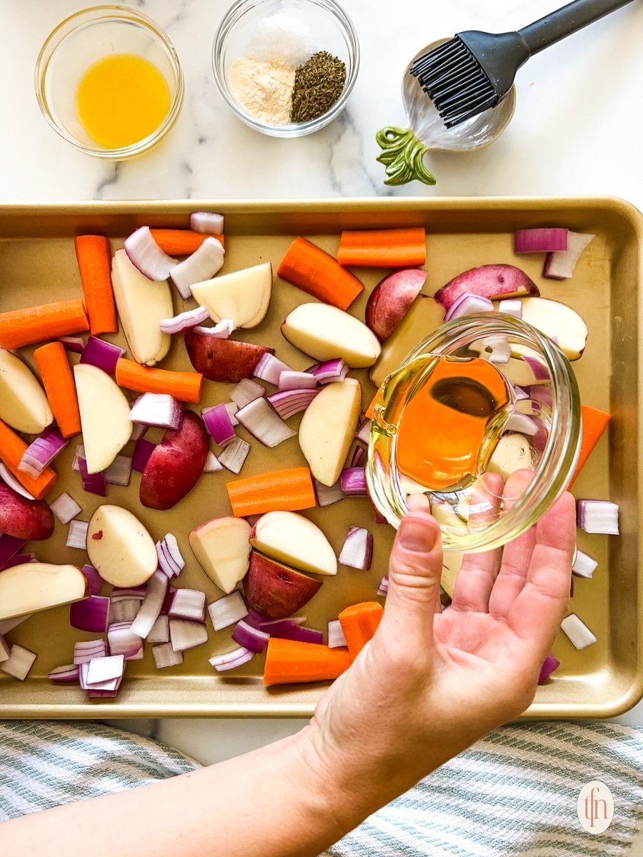 Hand drizzling oil onto chopped veggies laid out on a sheet pan.