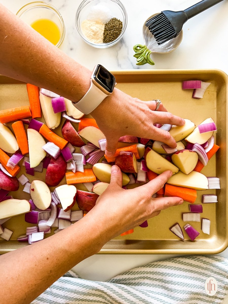 Hands adding chopped vegetables to a cooking sheet.