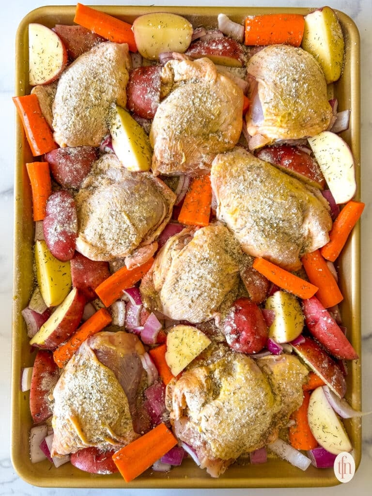 Jelly roll pan filled with seasoned, raw chicken thighs and chopped vegetables.