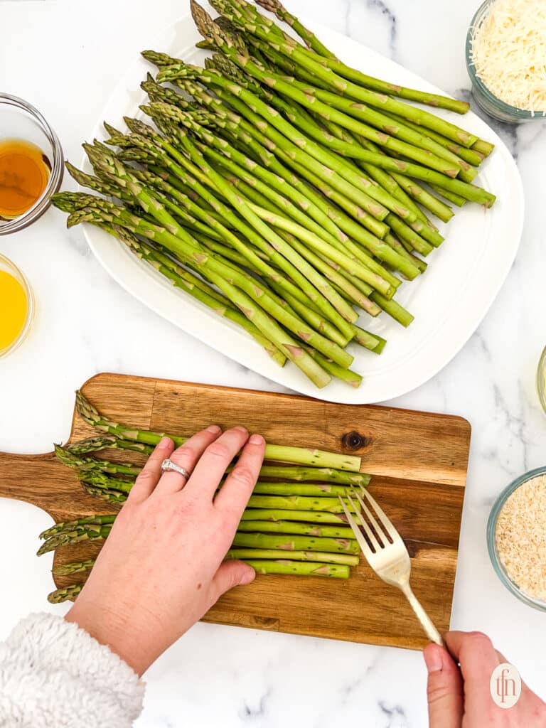 Poking asparagus using fork on top of a wooden chopping board with a plate of asparagus and a couple of ingredients.