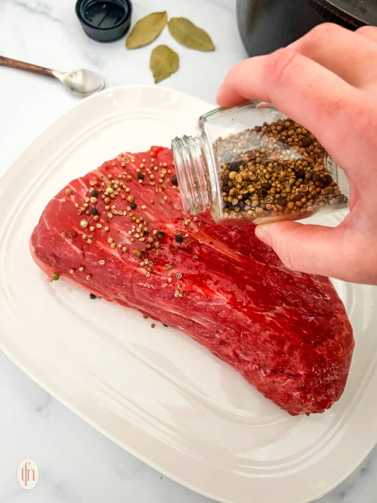 Seasoning being poured on top of raw beef placed on a white plate.