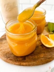 Two jars of meyer lemon curd with spoon on top of a wooden board with slices of lemon.