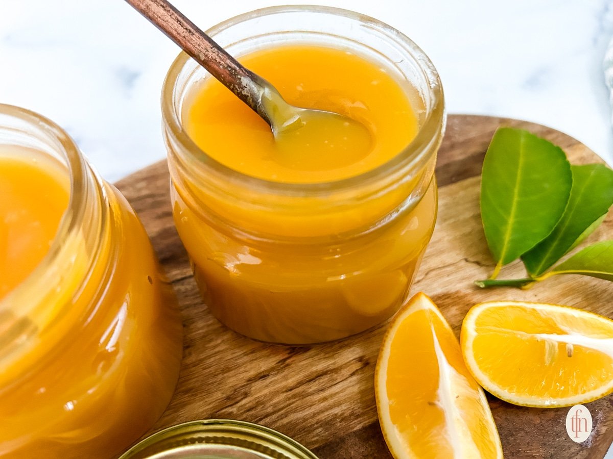 Two jars of meyer lemon curd with spoon on top of a wooden board with slices of lemon.