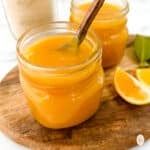 Two jars of meyer lemon curd with spoon on top of a wooden board with slices of lemon.