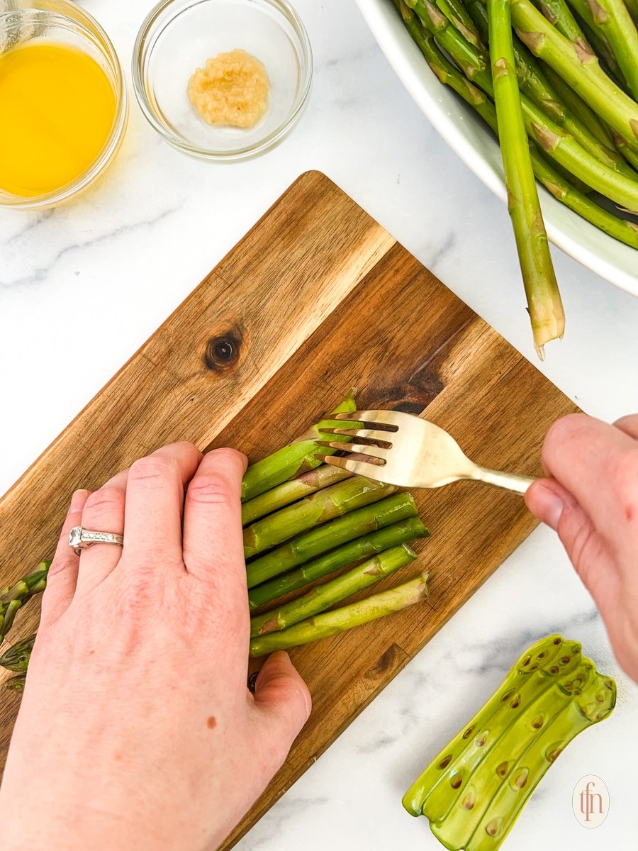 Poking the asparagus using fork on top of a wooden board.