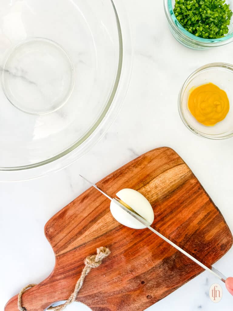 Egg being sliced on a wooden chopping board with a glass mixing bowl and a couple of prepared ingredients.
