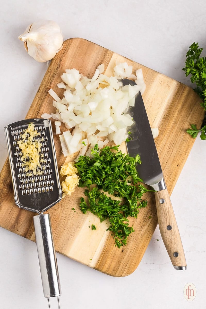 cutting board with knife, chopped parsley and onions, with grated garlic.