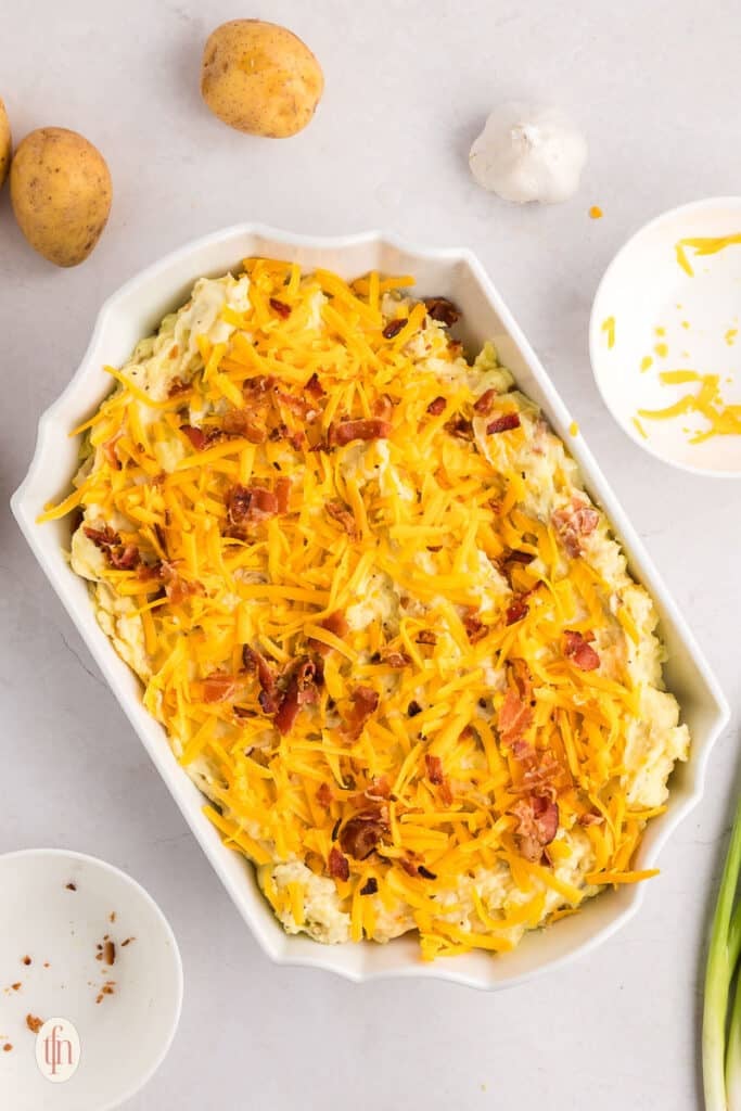 A mixture of mashed potatoes in a white casserole dish topped with bacon and cheddar cheese for twice baked mashed potatoes.