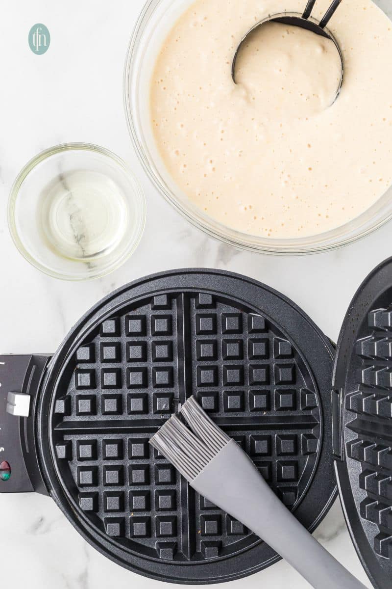 A waffle maker with a pastry brush and a bowl of batter mixture.