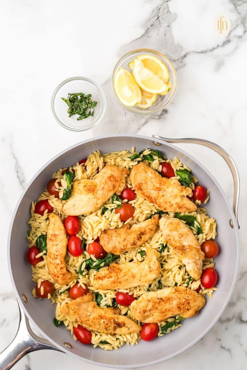A pan of chicken and orzo with a bowl of garnish and lemon.