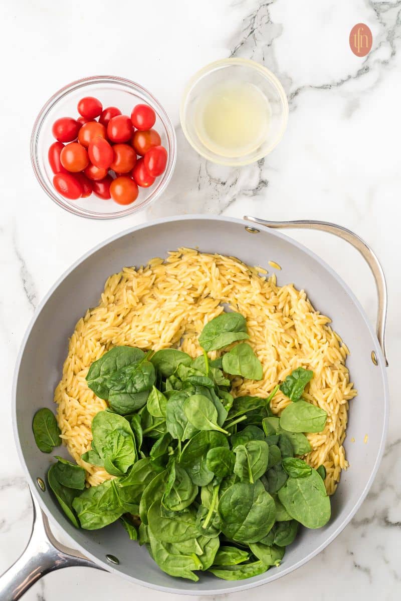 A pan of orzo with baby spinach and a bowl of liquid and tomatoes.
