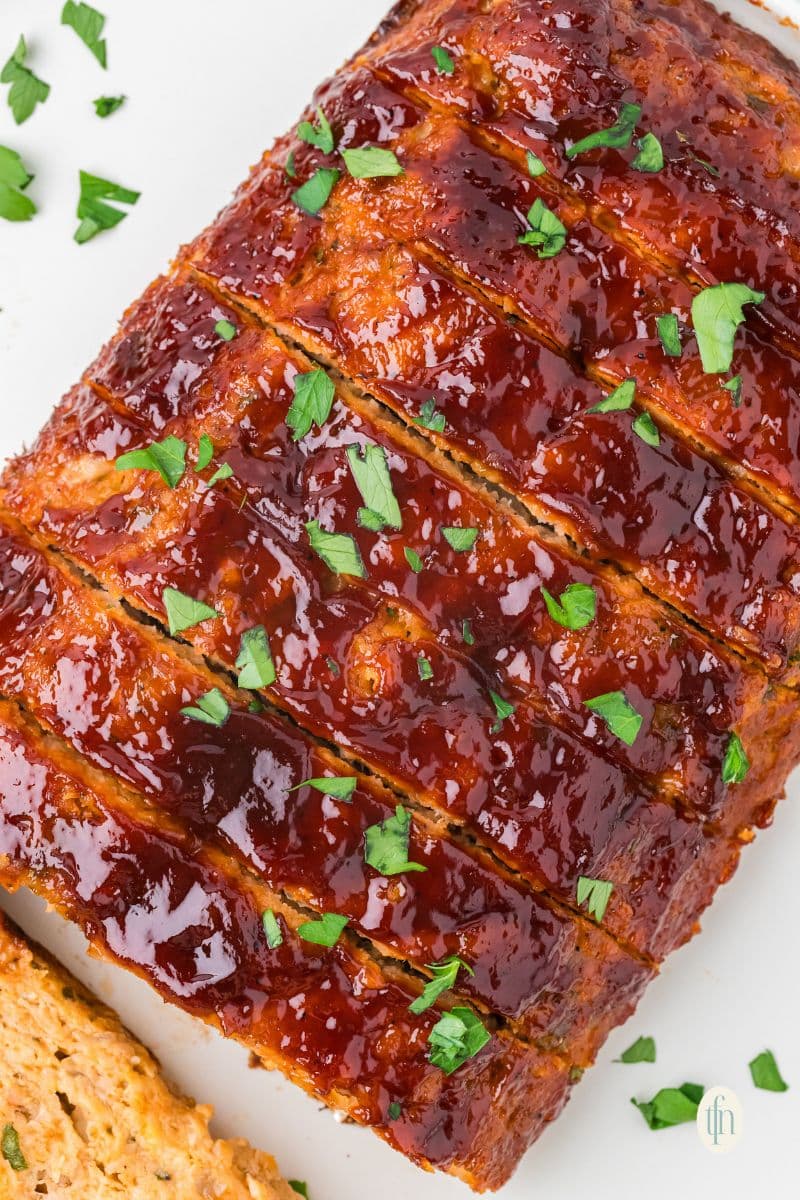 A close-up, overhead view of a sliced chicken meatloaf. The top is covered in a thick, shiny red glaze and sprinkled with chopped parsley.