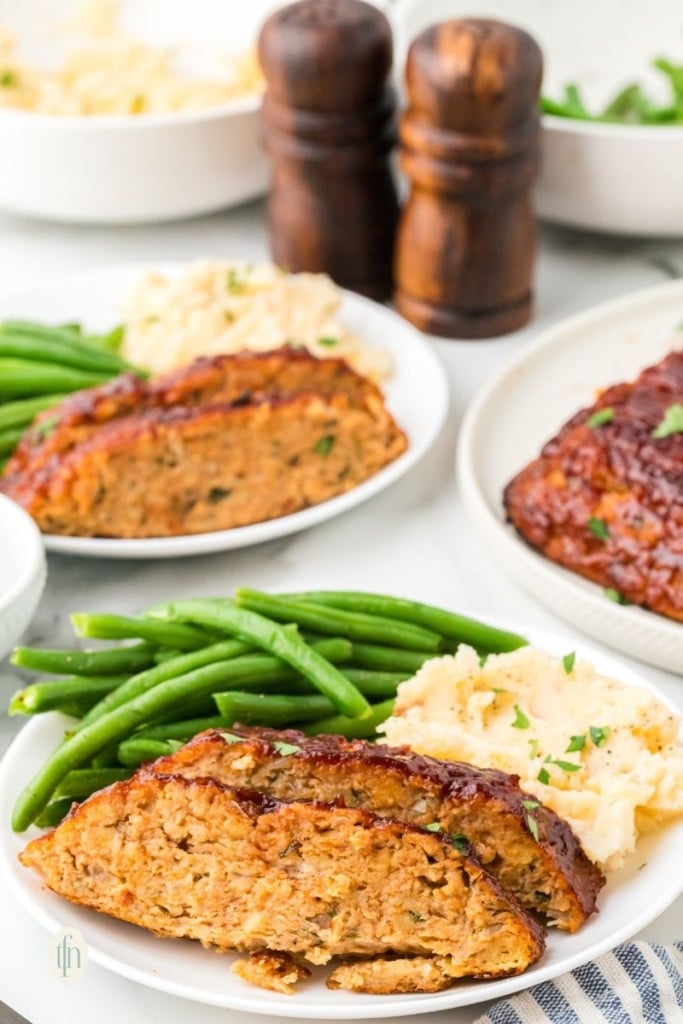 An eye-level shot showing two plates of glazed chicken meatloaf, mashed potatoes, and green beans, ready to be served. Two wooden spice shakers are visible in the background.