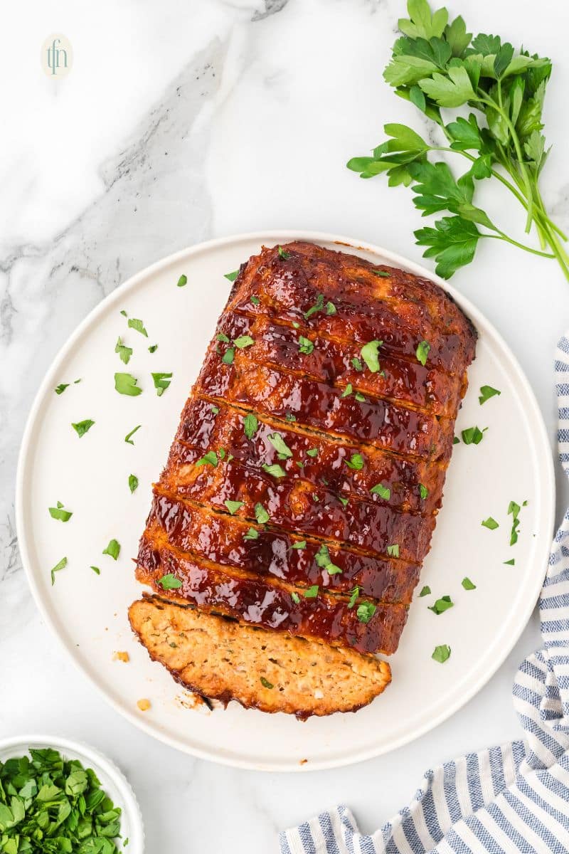 An overhead shot of a glazed chicken meatloaf on a white platter, already sliced into about eight thick pieces and garnished with chopped parsley.