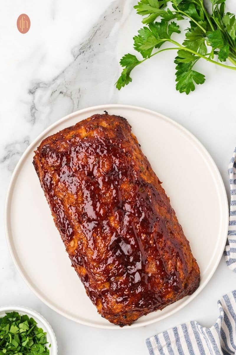 An overhead shot of a whole chicken meatloaf on a white platter. It is covered in a glossy, dark red glaze. A sprig of parsley and a blue and white striped towel are nearby.