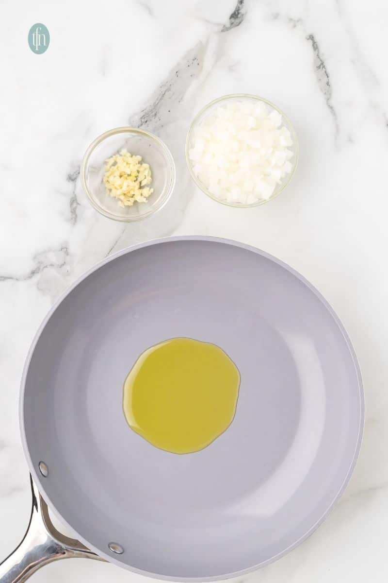 An overhead shot of a gray nonstick skillet with a pool of olive oil. Small glass bowls of chopped garlic and diced onion are visible on the marble counter.