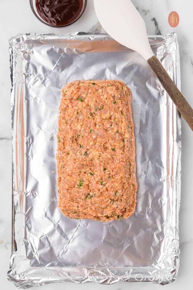 An overhead shot of the raw chicken meatloaf mixture shaped into a loaf on a foil-lined baking sheet. A white spatula is resting next to it, and a small bowl of glaze is visible at the top.
