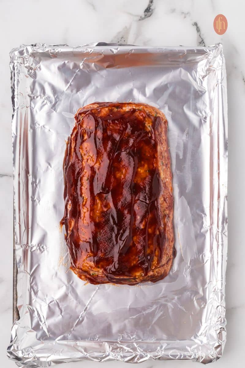 An overhead shot of the raw chicken meatloaf on a foil-lined baking sheet, now fully brushed with a generous layer of thick glaze before baking.