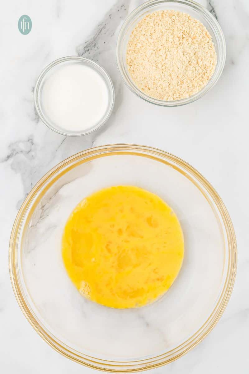 A large glass mixing bowl contains whisked eggs. Two small bowls are above it, one with milk and another with breadcrumbs.