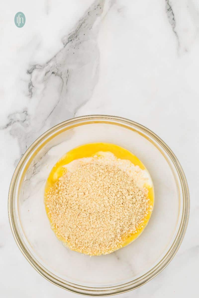 An overhead shot of a large glass bowl containing whisked eggs and milk, topped with a layer of dry breadcrumbs.