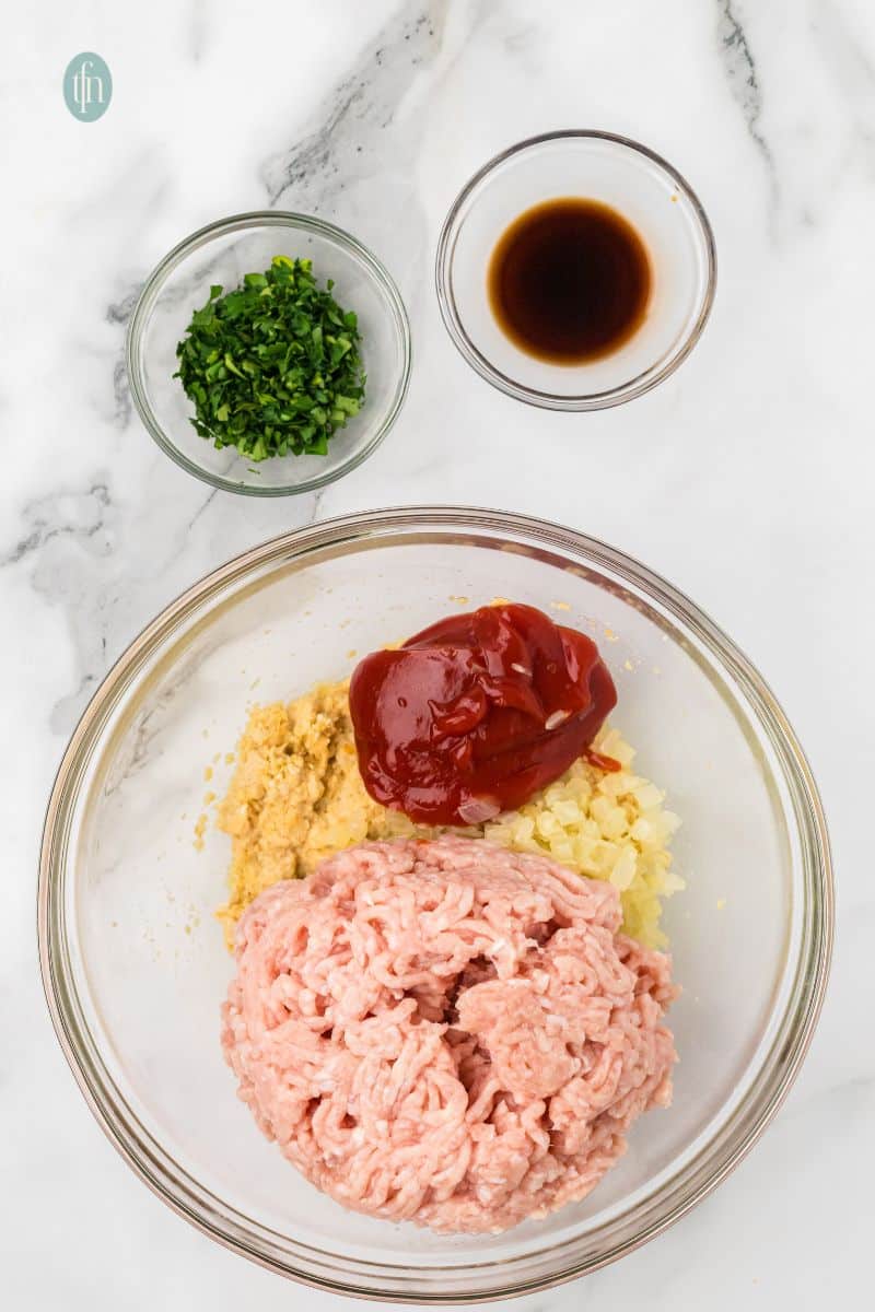 A large glass bowl is filled with raw ground chicken, the soaked breadcrumb mixture, sautéed onions/garlic, a dollop of ketchup, and a small amount of dark liquid (likely Worcestershire or soy sauce). A bowl of chopped parsley is on the side.