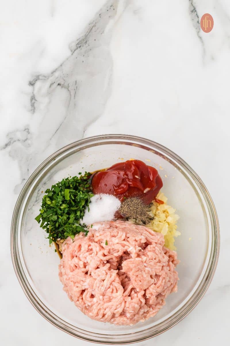 A close-up, overhead shot of the chicken meatloaf ingredients in the mixing bowl before being combined, showing the ground chicken, parsley, seasoning, and sauce components.