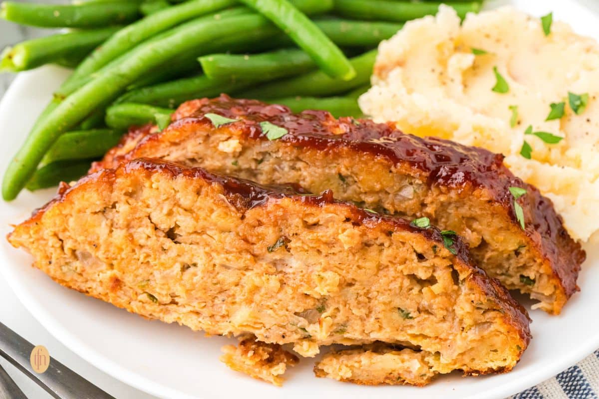 A wide, close-up shot of two slices of glazed chicken meatloaf served on a white plate with green beans and mashed potatoes.