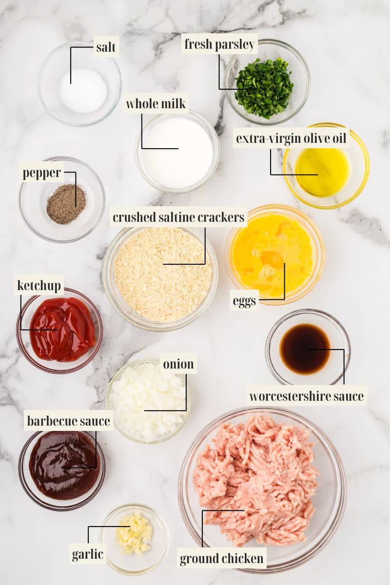 A flat lay of the labeled ingredients for ground chicken meatloaf: salt, pepper, ketchup, barbecue sauce, garlic, whole milk, crushed saltine crackers, diced onion, ground chicken, fresh parsley, extra virgin olive oil, eggs, and Worcestershire sauce.