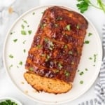 A square, overhead shot of a whole chicken meatloaf on a round white platter. It is already sliced and topped with a glossy glaze and chopped parsley.