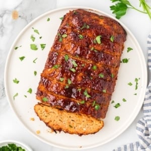 A square, overhead shot of a whole chicken meatloaf on a round white platter. It is already sliced and topped with a glossy glaze and chopped parsley.