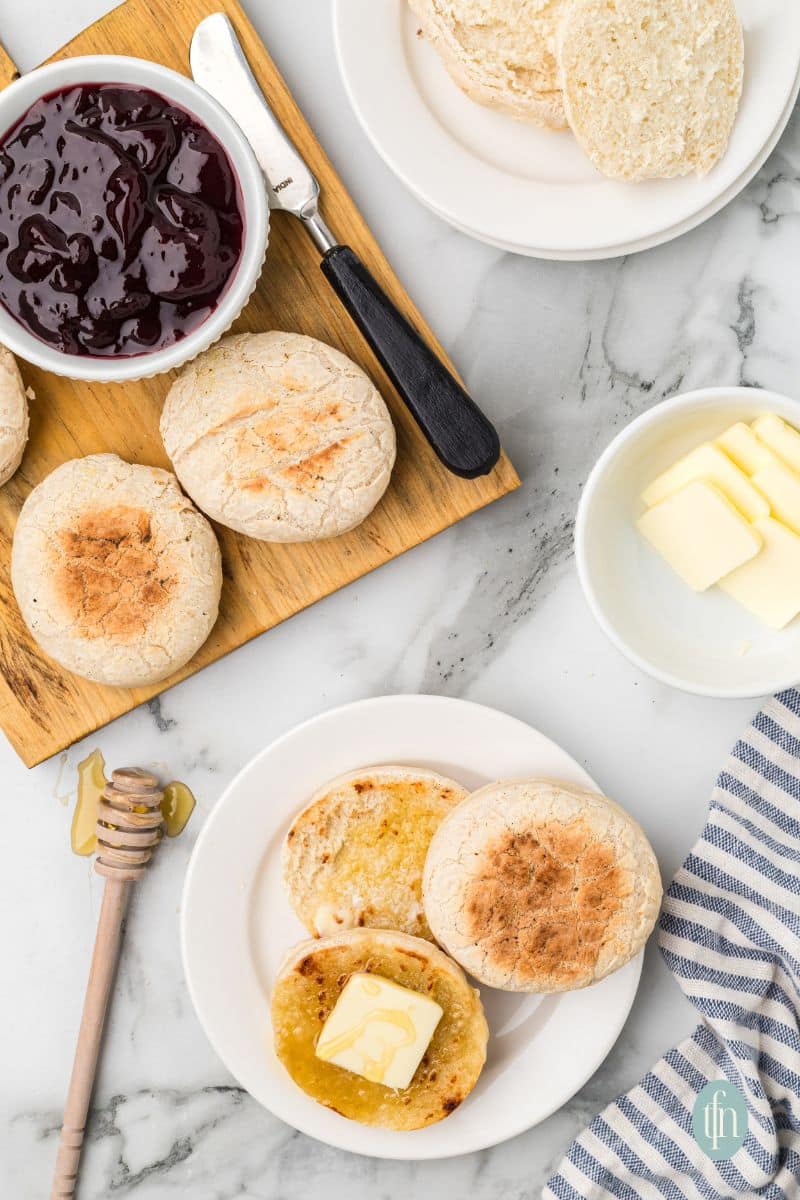 A plate of english muffin with butter on top and a wooden platter of muffins with a bowl of jam.